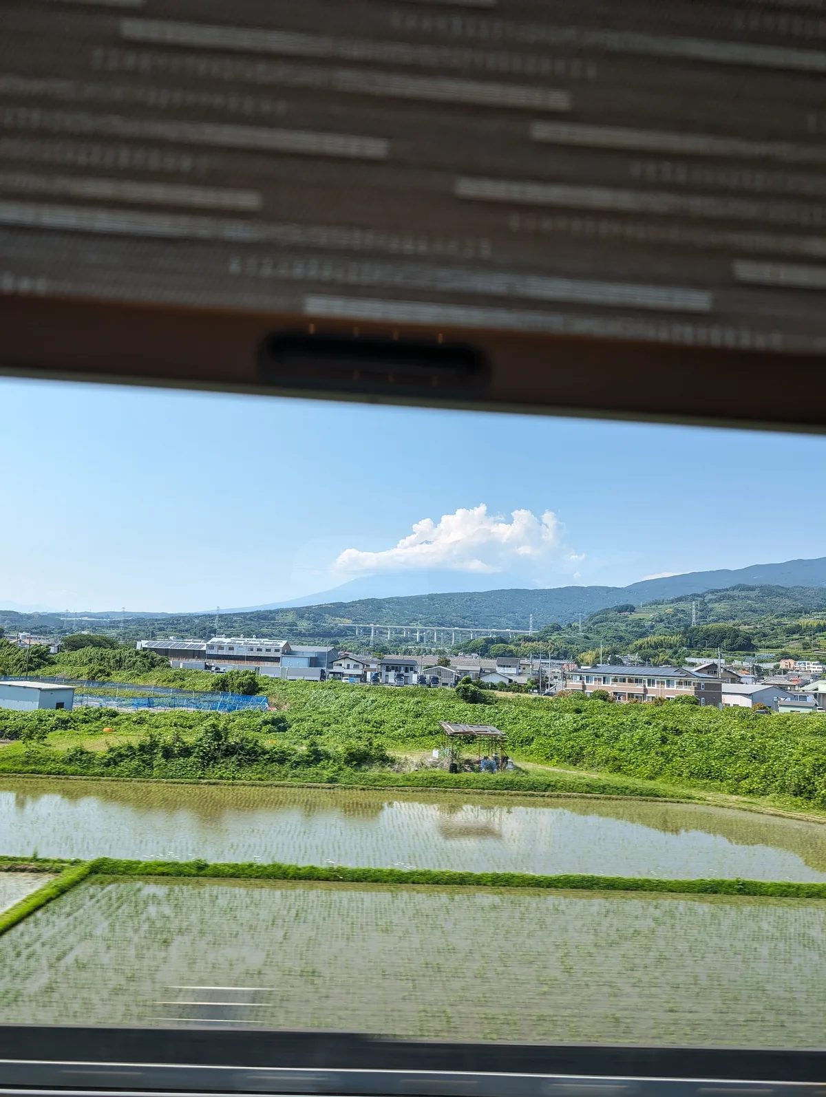 Scenic view from a Shinkansen train window of green rice paddies, scattered houses, and distant mountains in Japan.