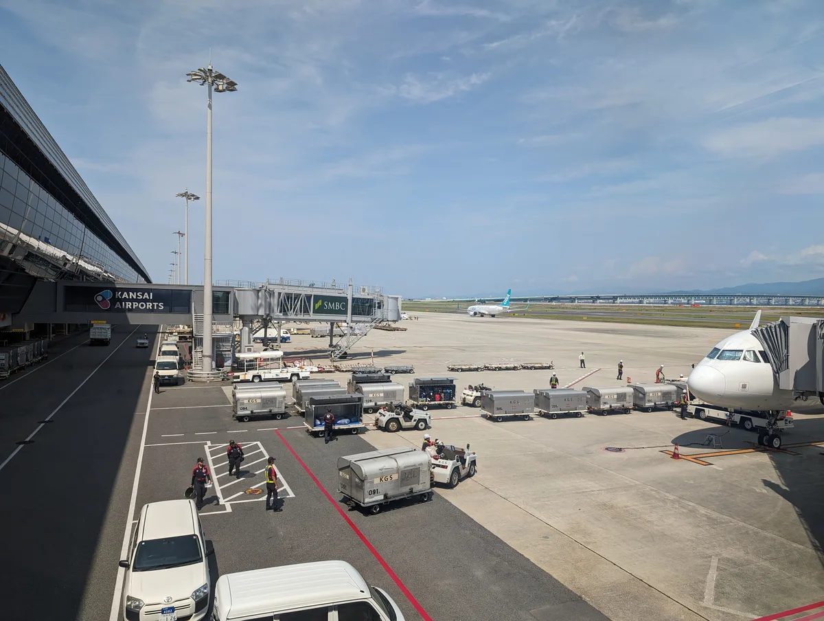 Kansai International Airport tarmac with jet bridge, ground crew, and planes under a partly cloudy sky in Osaka, Japan.