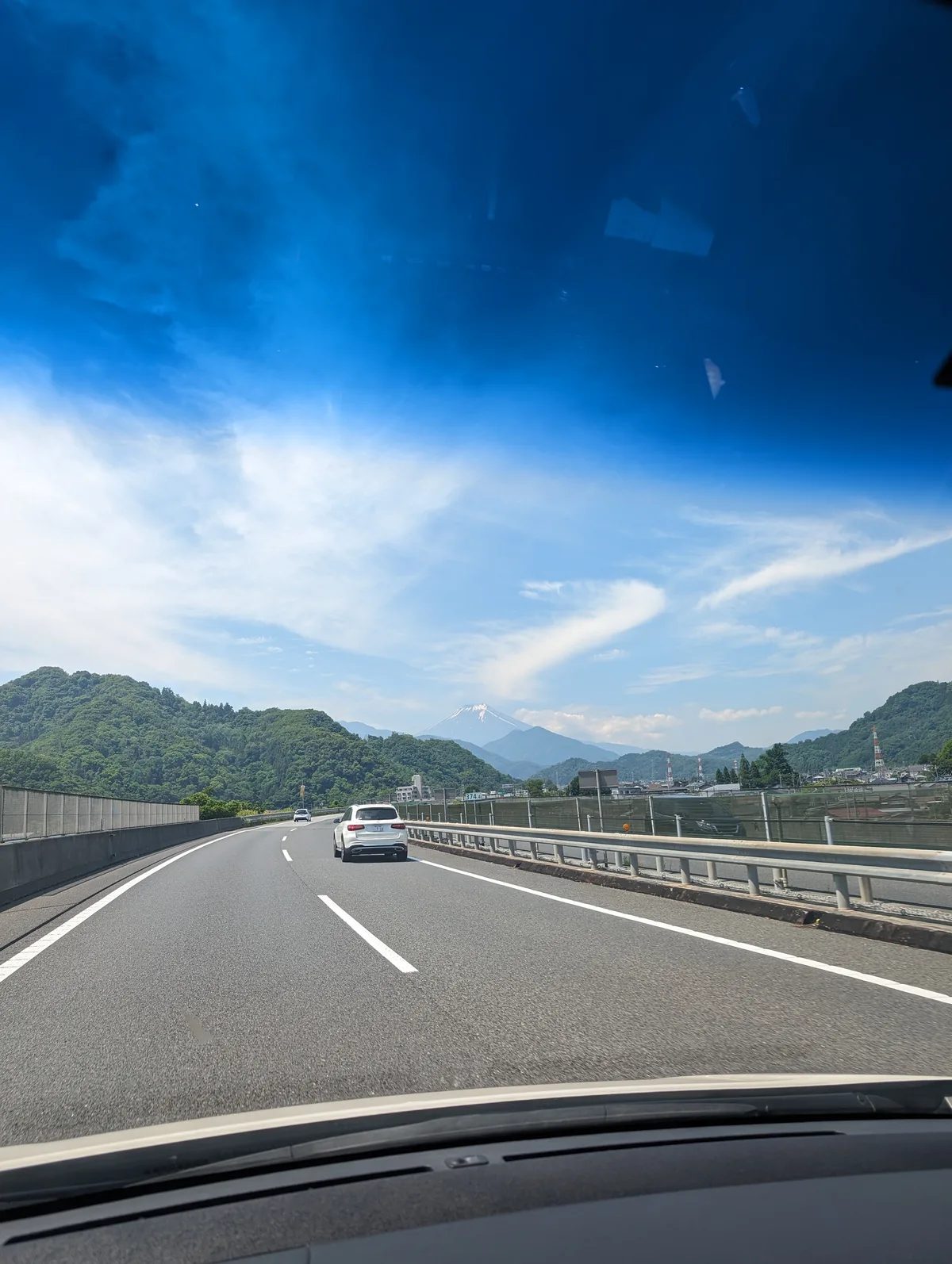 Mount Fuji in the distance from a highway in Japan, flanked by lush green mountains under a bright blue sky.