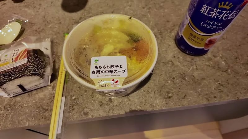 Overhead shot of a Family Mart convenience store meal — glass noodle soup, dumplings, rice ball, and milk tea on a grey counter in Tokyo, Japan