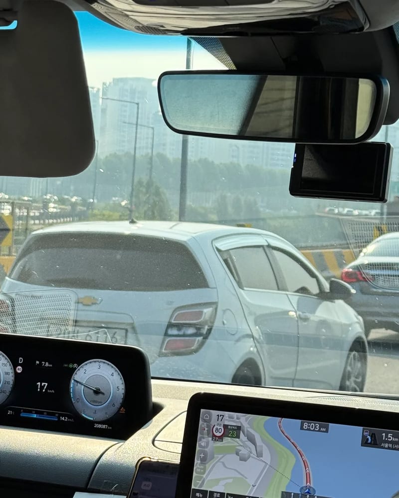 View through the windshield of the Staria on the highway into Seoul with a white Chevrolet hatchback alongside and a navigation screen on the dash