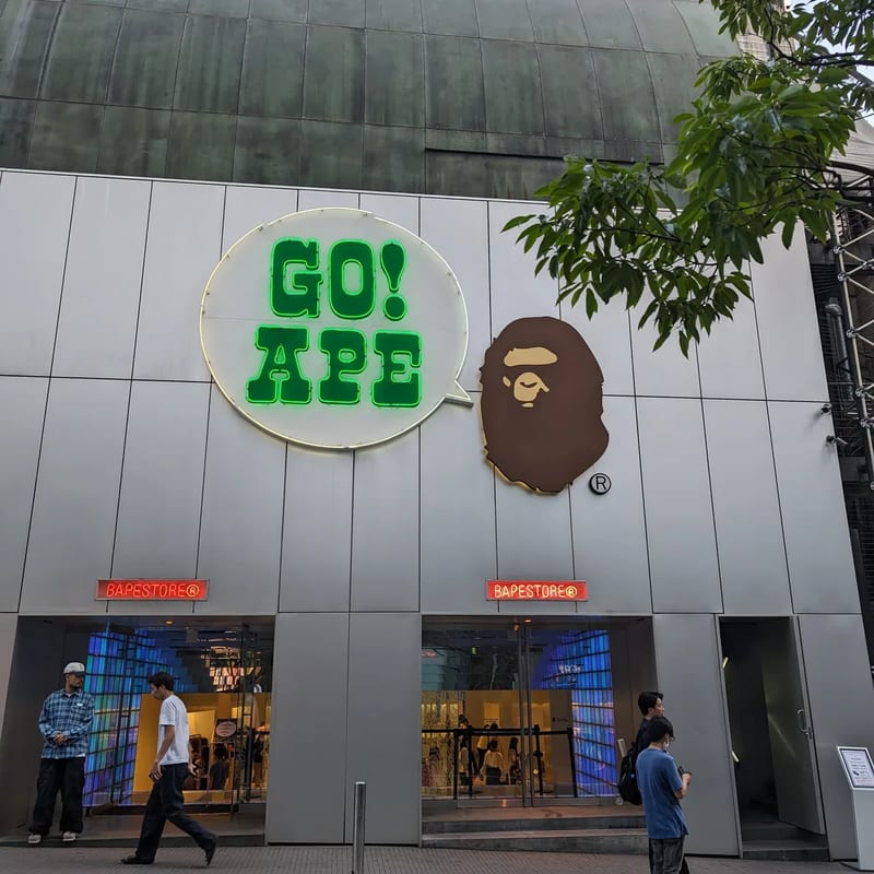 BAPE store exterior in Shibuya, Tokyo, Japan, with 'GO! APE' neon sign and ape head logo, pedestrians walking past.