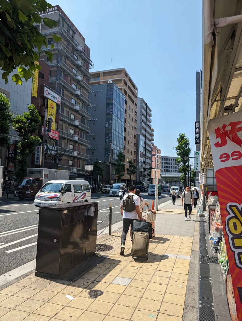 Person pulling luggage on a yellow-tiled sidewalk in Osaka, Japan, next to a busy road under a clear sky.
