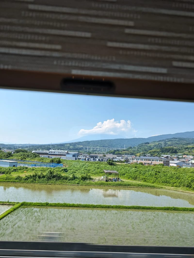 Scenic view from a Shinkansen train window of green rice paddies, scattered houses, and distant mountains in Japan.