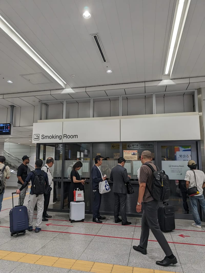 Glass-enclosed smoking room in Shin-Osaka train station with travelers and luggage, Japan.