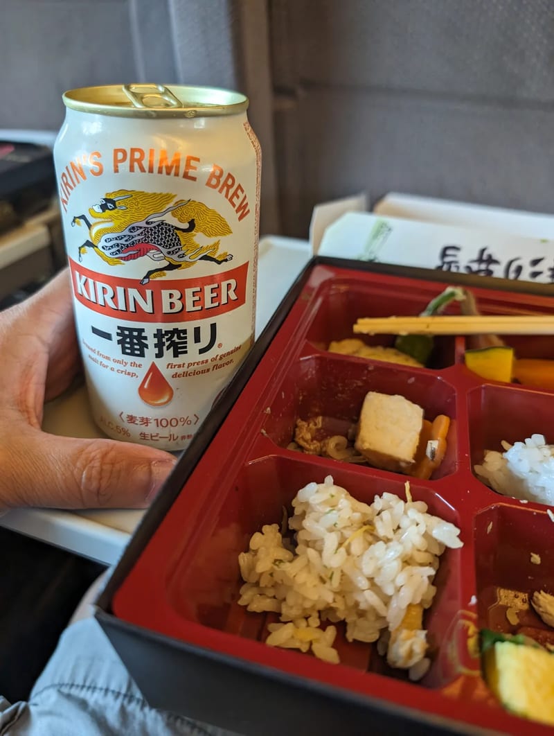 Hand holding a Kirin beer can next to an open Japanese bento box on a Shinkansen tray table, Japan.