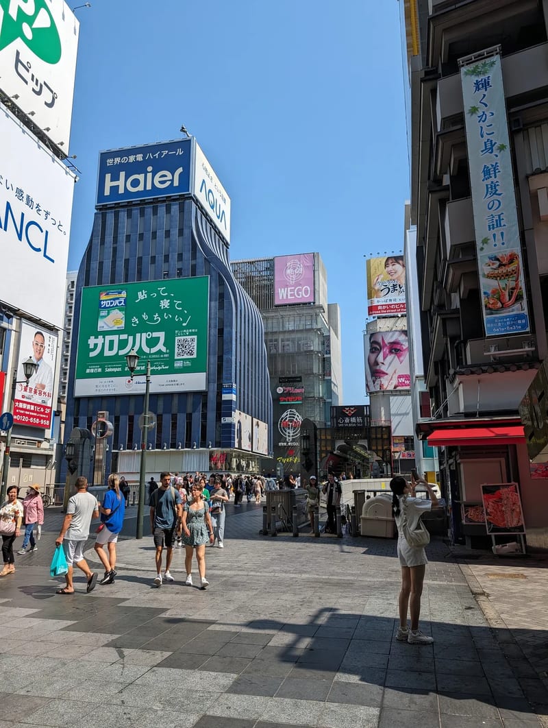 Wide pedestrian street in Osaka, Japan, filled with people between tall buildings and large electronic billboards.