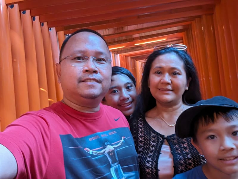 Family selfie inside Fushimi Inari Shrine's orange torii gate tunnel in Kyoto, Japan.