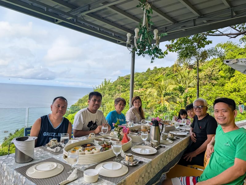 Family group seated at an outdoor dining table with a panoramic ocean and green hill view at Casa Amara, Batangas, Philippines.