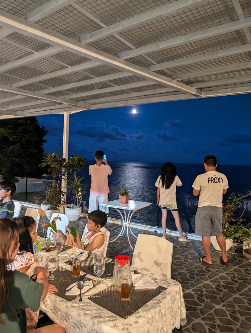 People at the edge of an outdoor patio photographing a full moon reflecting brightly on the dark ocean at night at Casa Amara, Batangas, Philippines.