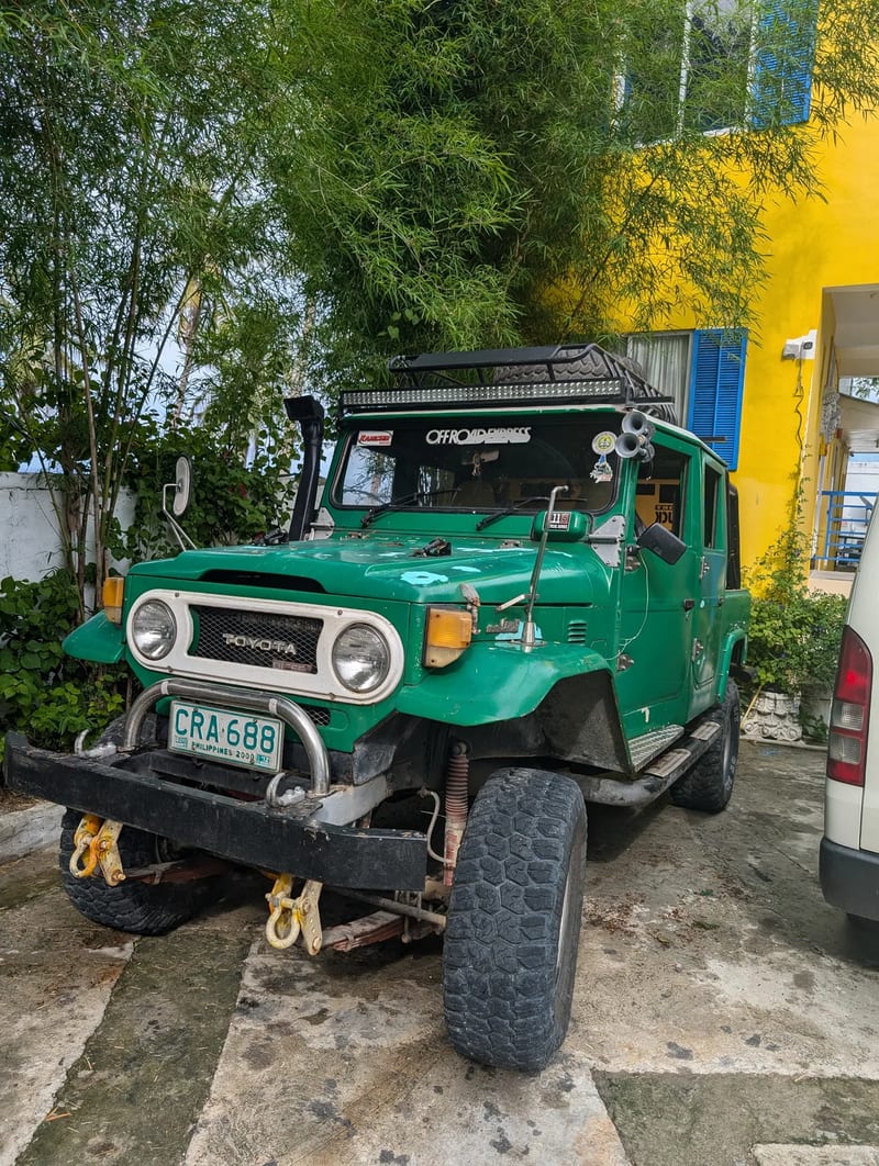 Bright green vintage Toyota Land Cruiser with roof rack, snorkel, and large off-road tires parked in front of a yellow building with bamboo at Casa Amara, Batangas, Philippines.