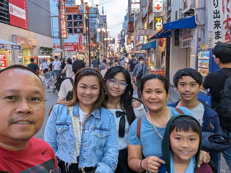 Family selfie smiling in a busy Dotonbori street in Osaka, Japan with neon signs in the background.