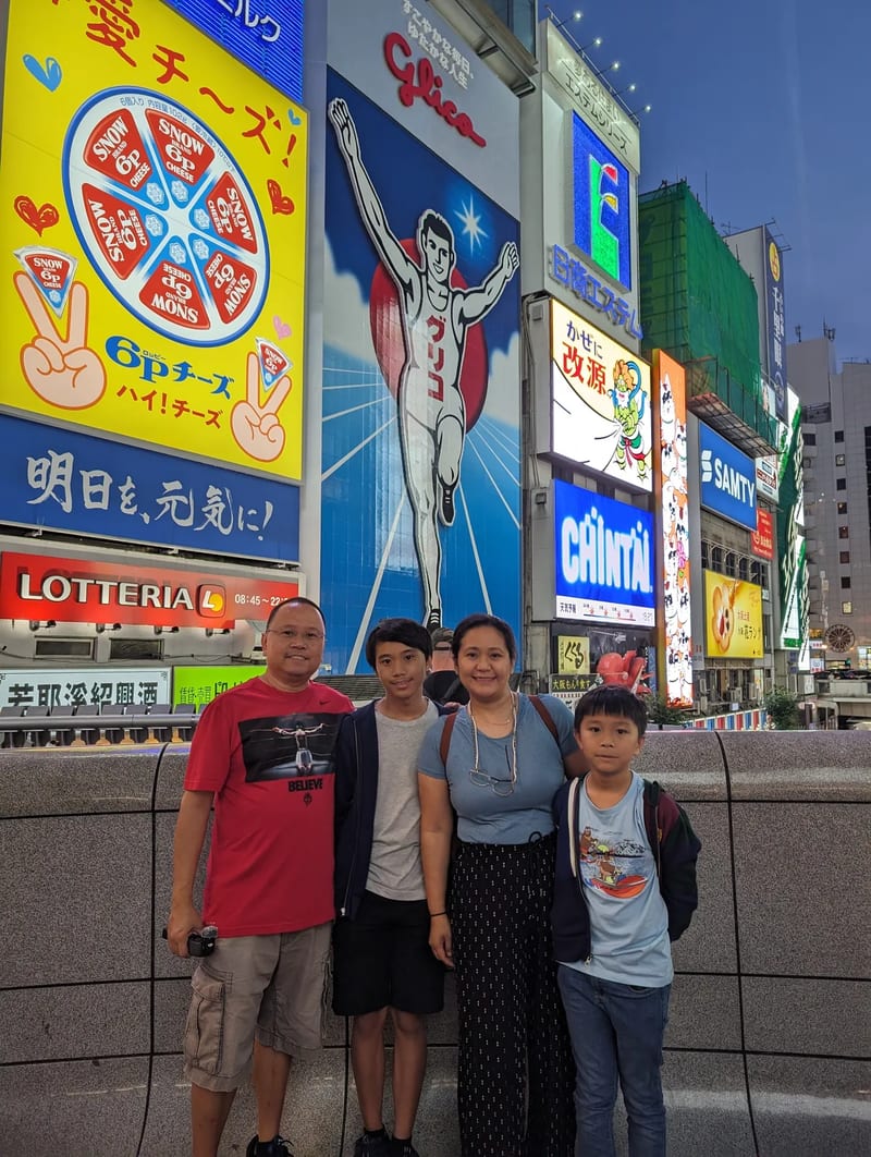 Family of four in front of the iconic Glico Man sign and neon billboards in Dotonbori, Osaka, Japan.