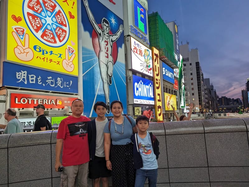 Family of four posing in front of the Glico Man sign with a wide view showing the Dotonbori skyline, Osaka, Japan.