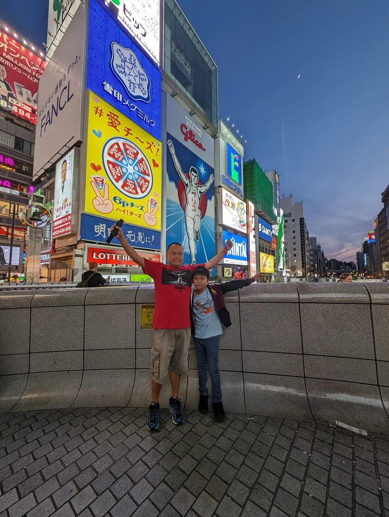 Father and son with arms raised mimicking the iconic Glico Man pose in front of the Glico Man sign in Dotonbori, Osaka, Japan.