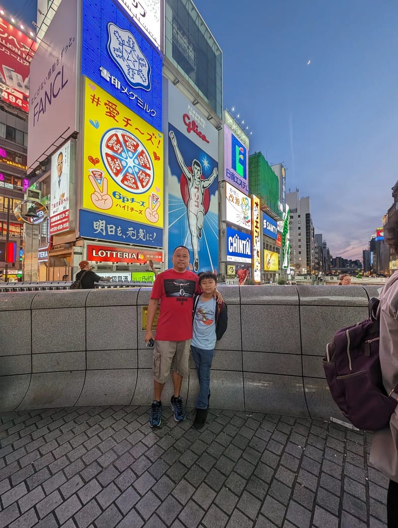 Father and son posing in front of the iconic Glico Man sign with neon billboards in Dotonbori, Osaka, Japan.