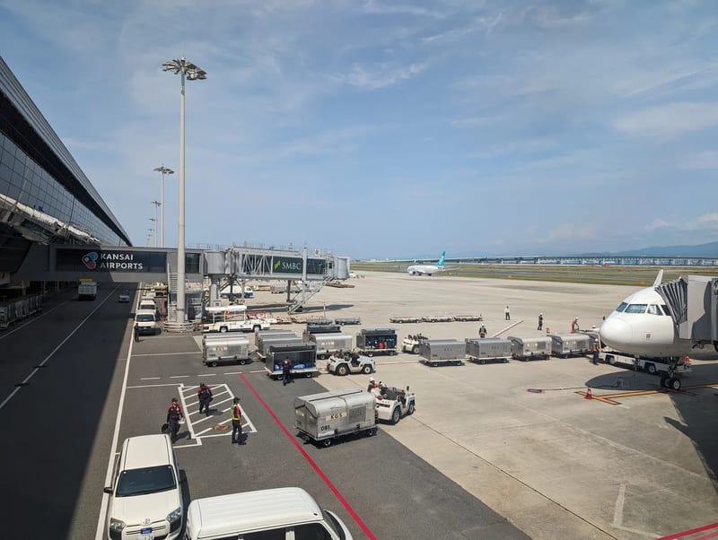 Kansai International Airport tarmac with jet bridge, ground crew, and planes under a partly cloudy sky in Osaka, Japan.