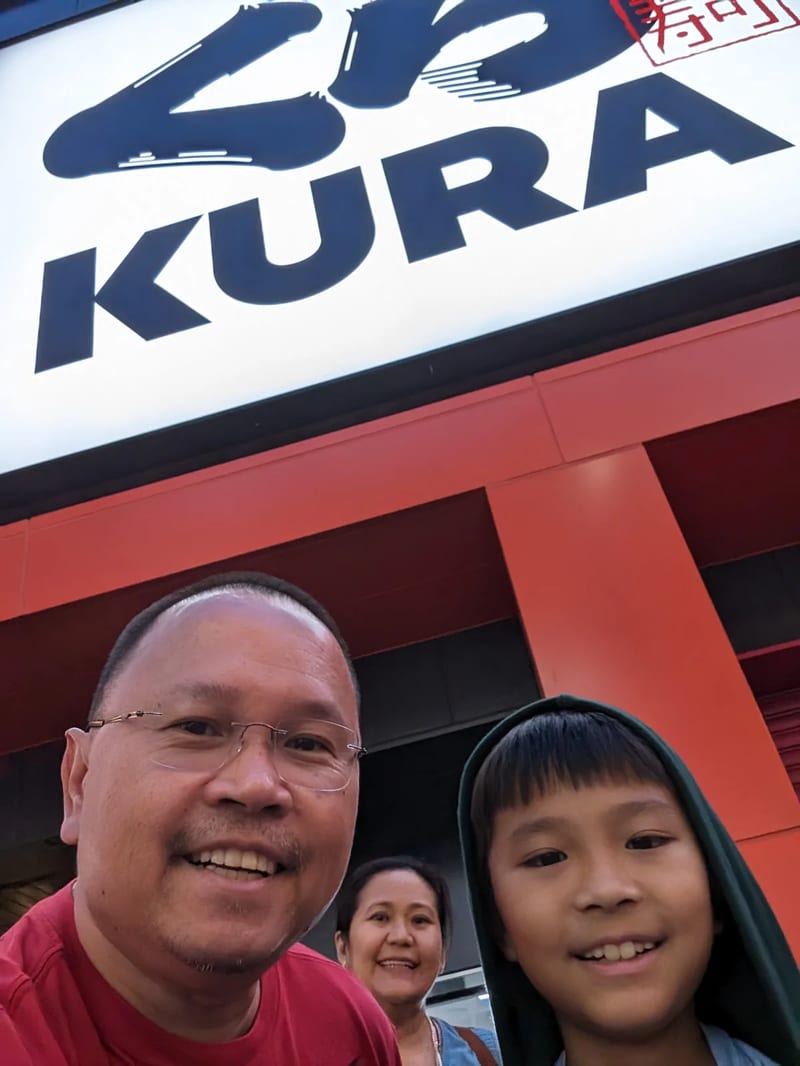 Man and boy selfie smiling in front of a large illuminated Kura Sushi sign in Osaka, Japan.