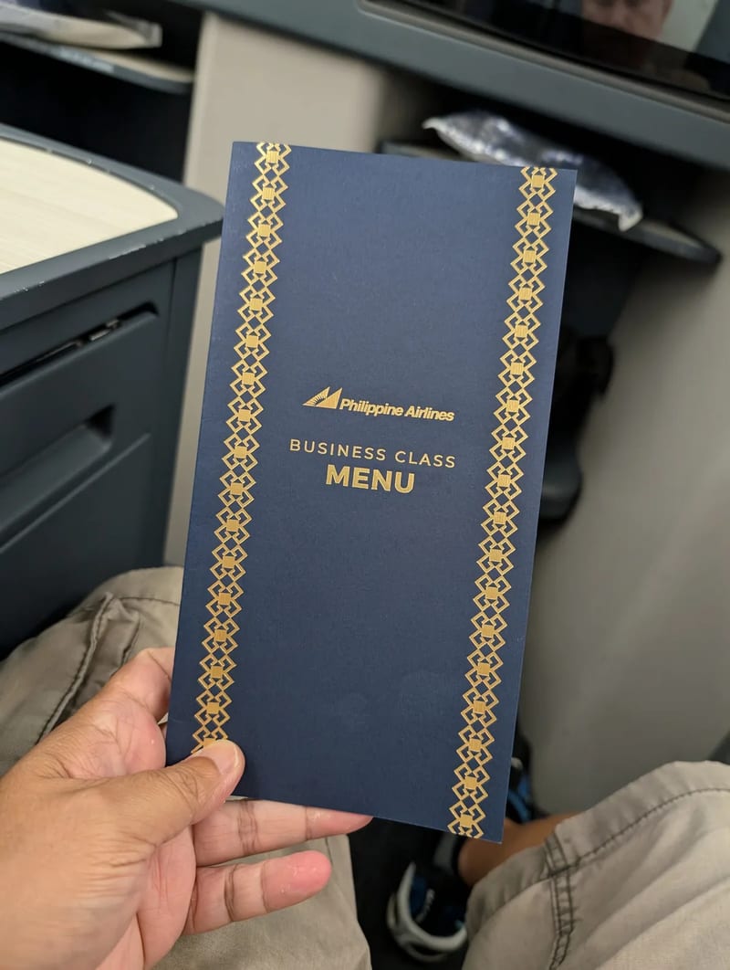 Hand holding a dark blue Philippine Airlines Business Class menu with gold patterned borders in the airplane cabin, Haneda Airport, Tokyo, Japan.