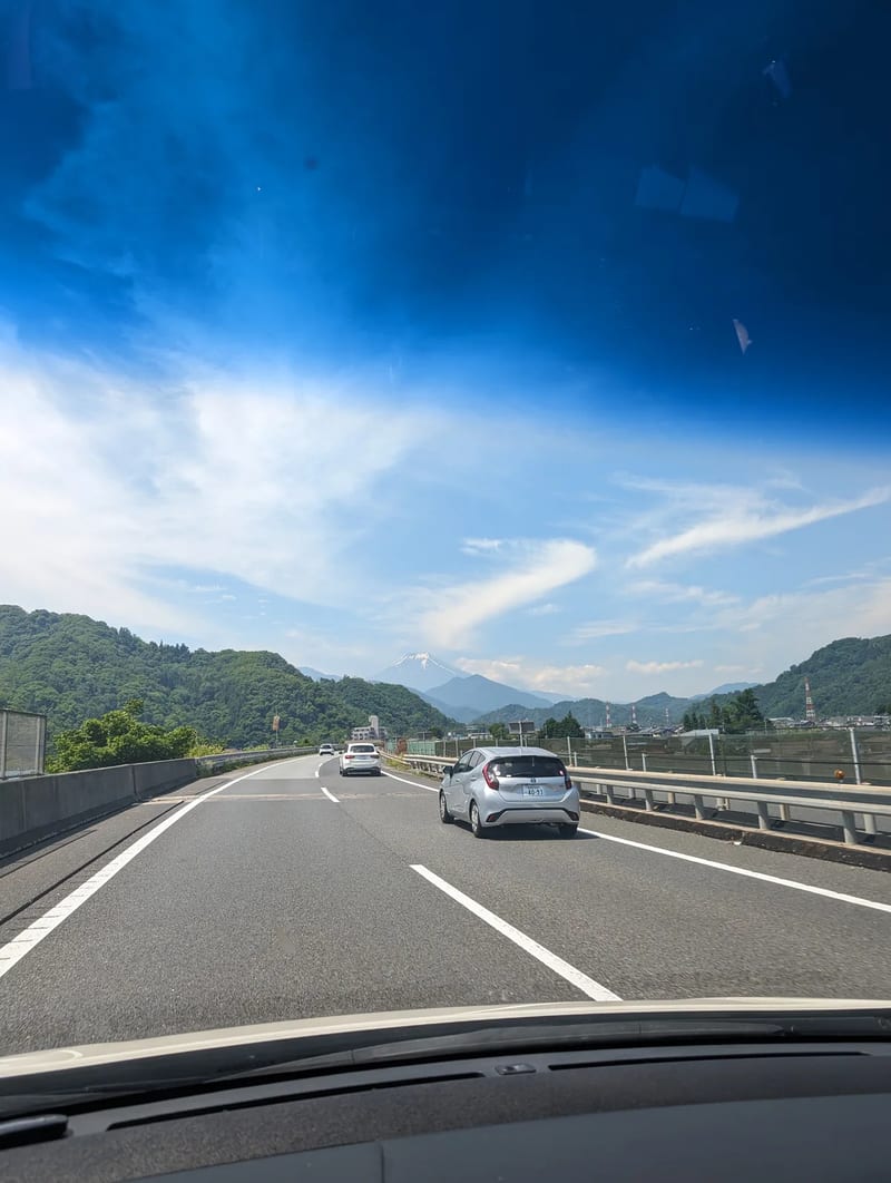 Mount Fuji visible from a highway in Yamanashi, Japan, silver car in foreground, blue sky.