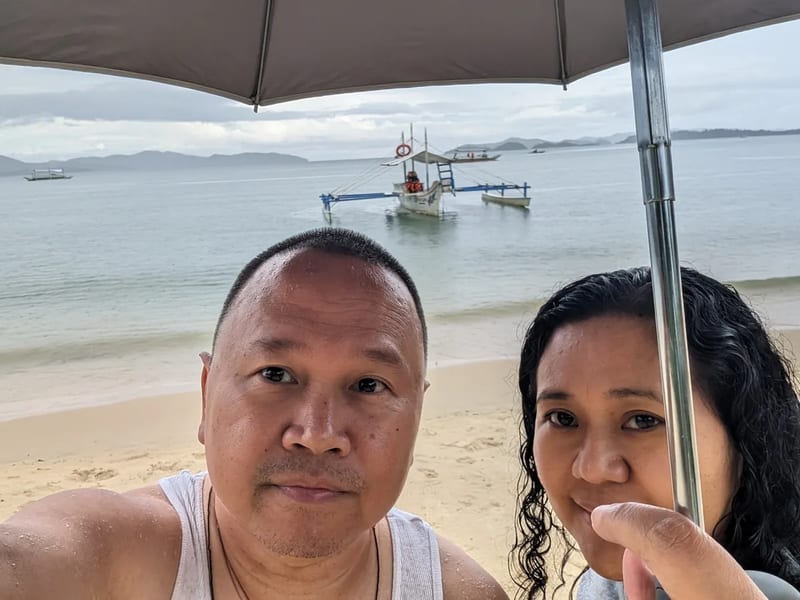 Close-up selfie of a couple under an umbrella on Port Barton beach Palawan with a traditional outrigger boat in the background