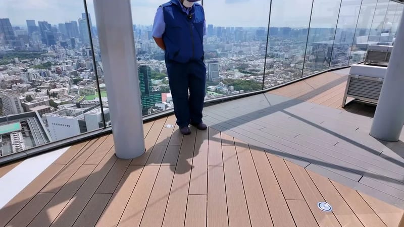 Staff member in blue uniform on the Shibuya Sky outdoor rooftop deck in Shibuya, Tokyo, Japan, city view and wooden flooring visible.