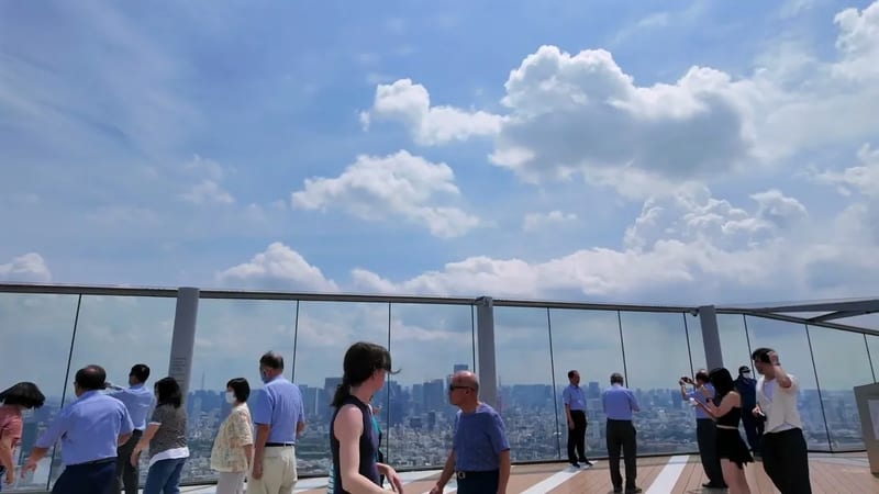 People on the outdoor rooftop observation deck of Shibuya Sky in Tokyo, Japan, with panoramic city views through clean glass barriers under a bright, cloudy sky.