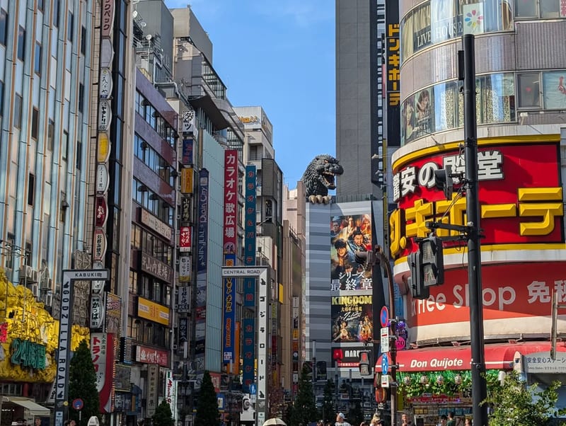 Bustling Shinjuku street in Tokyo, Japan, lined with tall buildings covered in vibrant neon signs and advertisements, with the iconic Godzilla Head visible peeking above a building in the distance.