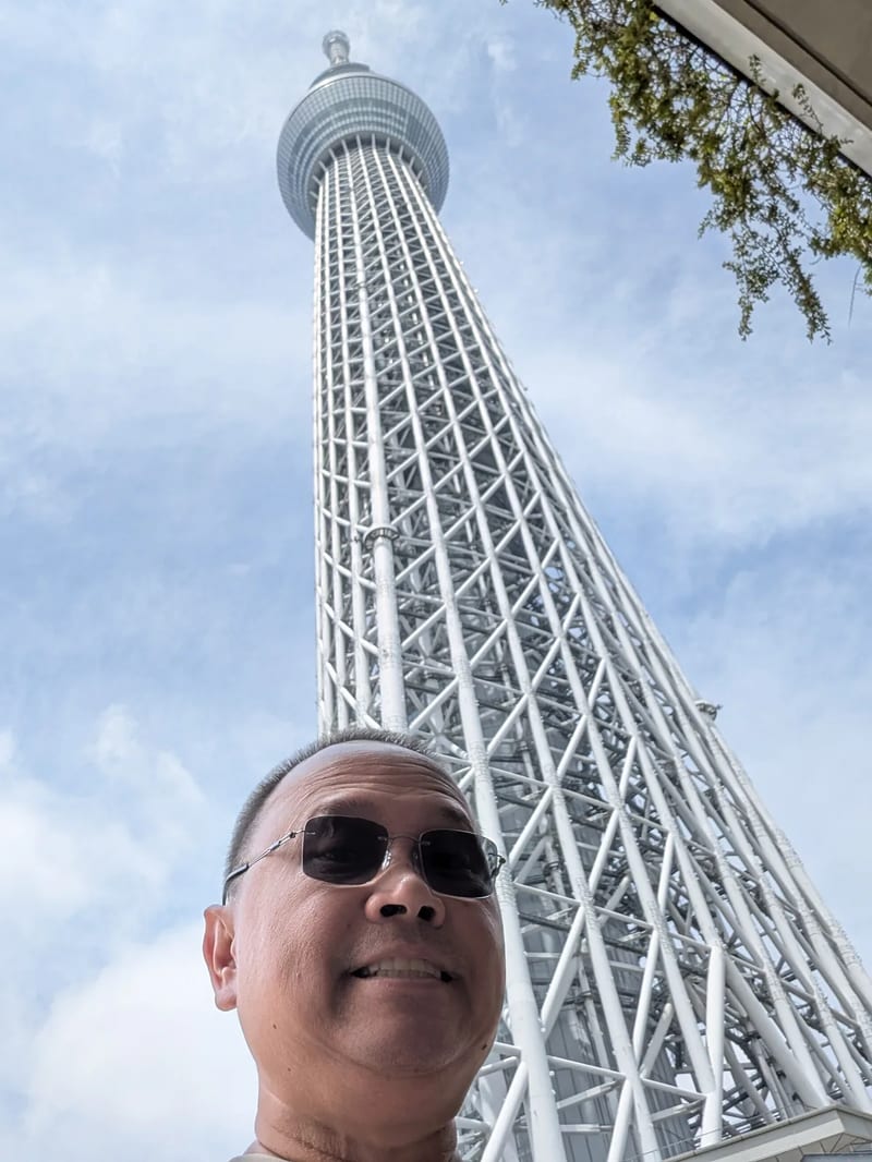 Man in sunglasses smiling at the camera with the Tokyo Skytree towering behind him against a blue sky with light clouds, Tokyo, Japan.